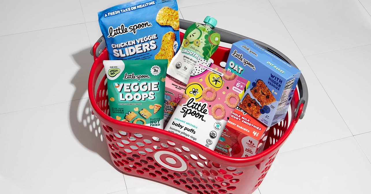 Red Target shopping basket filled with various Little Spoon kids’ food products, including Veggie Loops, Baby Puffs, Smoothies, Chicken Veggie Sliders, and Oat Bakes, placed on a white tiled floor