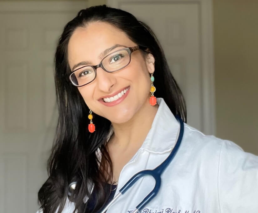 Dr. Krupa Playforth, also known as The Pediatrician Mom, smiling warmly in a white coat with a stethoscope draped over her shoulder, wearing glasses and colorful earrings.