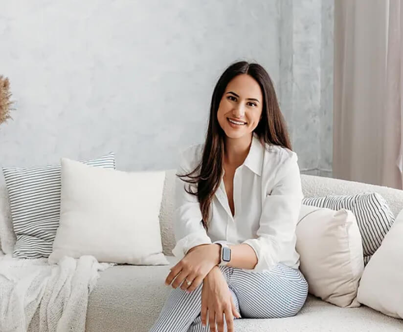 Dr. Sarah Habibi, Scientist and STEM Educator, seated on a cozy couch with pillows, smiling warmly while wearing a white shirt and striped pants in a bright, serene setting.