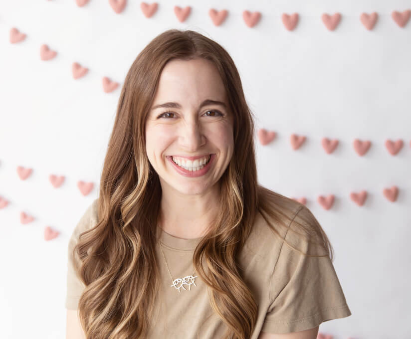 Dr. Taylor Arnold, pediatric dietitian, smiling brightly with long wavy hair, standing in front of a backdrop decorated with small pink heart-shaped garlands.