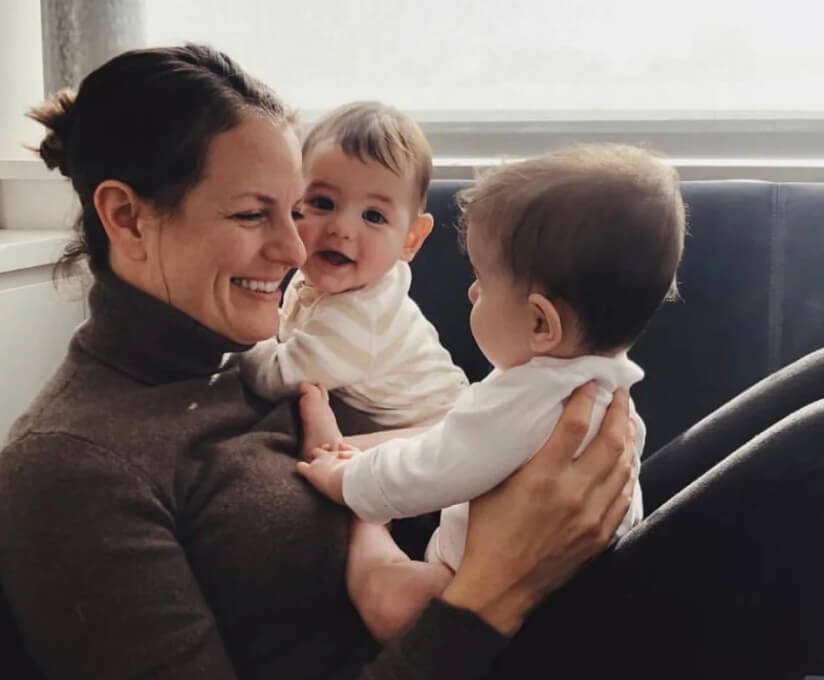 Jenny Best, Founder of Solid Starts, smiling and holding two babies in her arms while seated, with soft lighting from a nearby window.