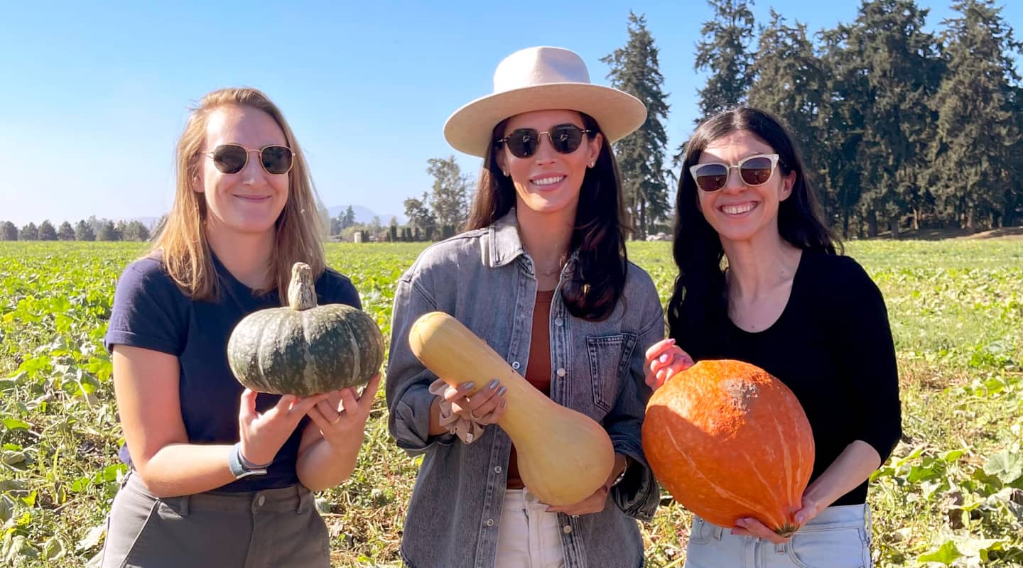 Three members of the Little Spoon team standing in a field at one of their farms, each holding a different type of squash. Rows of green crops stretch behind them as they smile and enjoy their time.