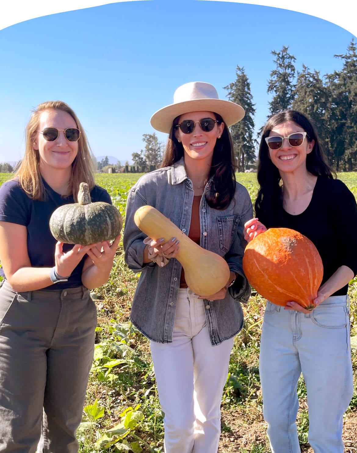 Three members of the Little Spoon team standing in a field at one of their farms, each holding a different type of squash. Rows of green crops stretch behind them as they smile and enjoy their time.