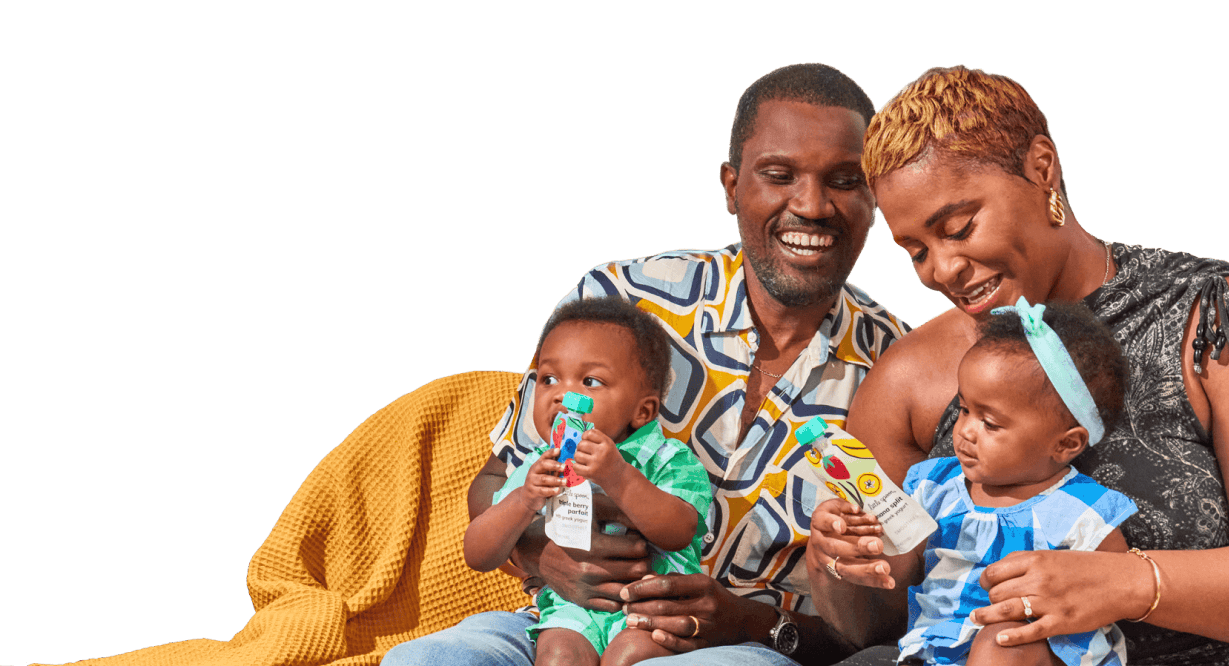 A smiling family of four, with a mother, father, and two young children sitting together on a couch. The parents hold their children, each holding a Little Spoon Smoothies pouch.