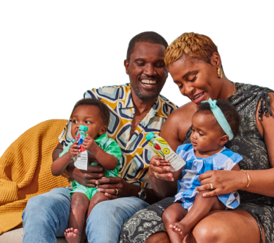 A smiling family of four, with a mother, father, and two young children sitting together on a couch. The parents hold their children, each holding a Little Spoon Smoothies pouch.