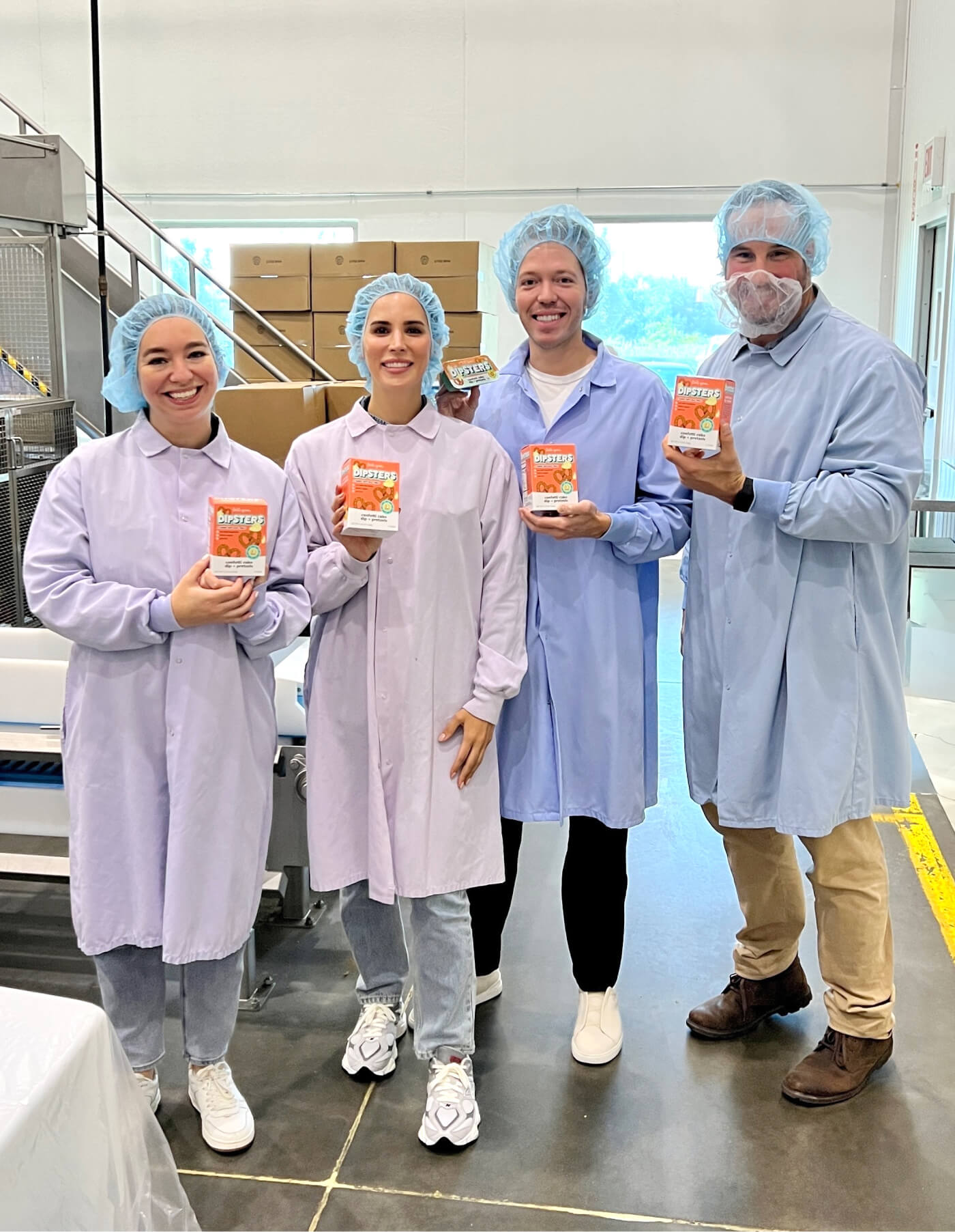 A group of four Little Spoon team members standing inside a facility, wearing hair nets and lab coats. They are smiling and holding up Little Spoon Dipsters, proudly showcasing the packaging.