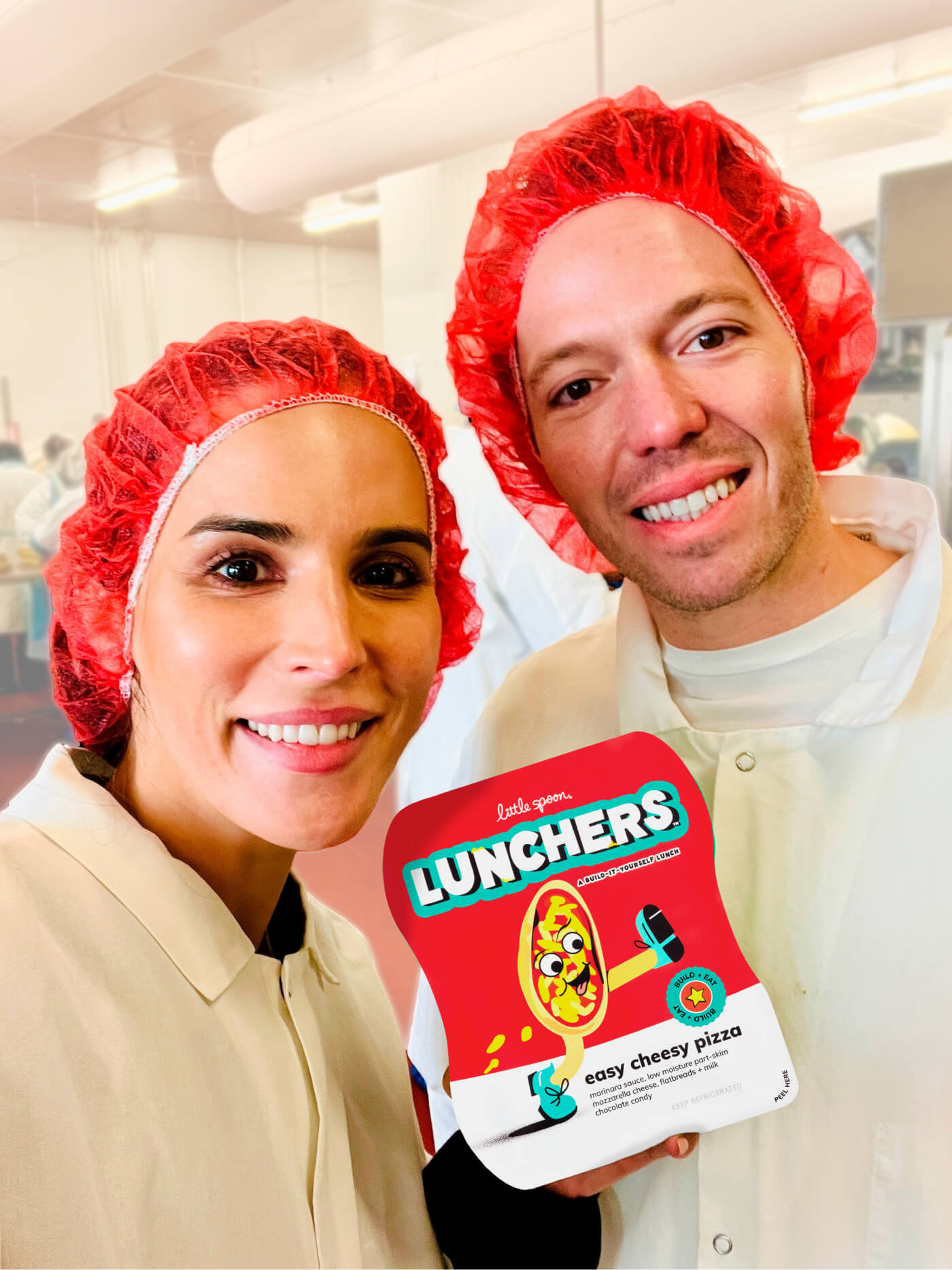 Little Spoon co-founders Ben and Angela, both wearing red hairnets and white lab coats, smiling and holding a Lunchers Easy Cheesy Pizza package inside a production facility.