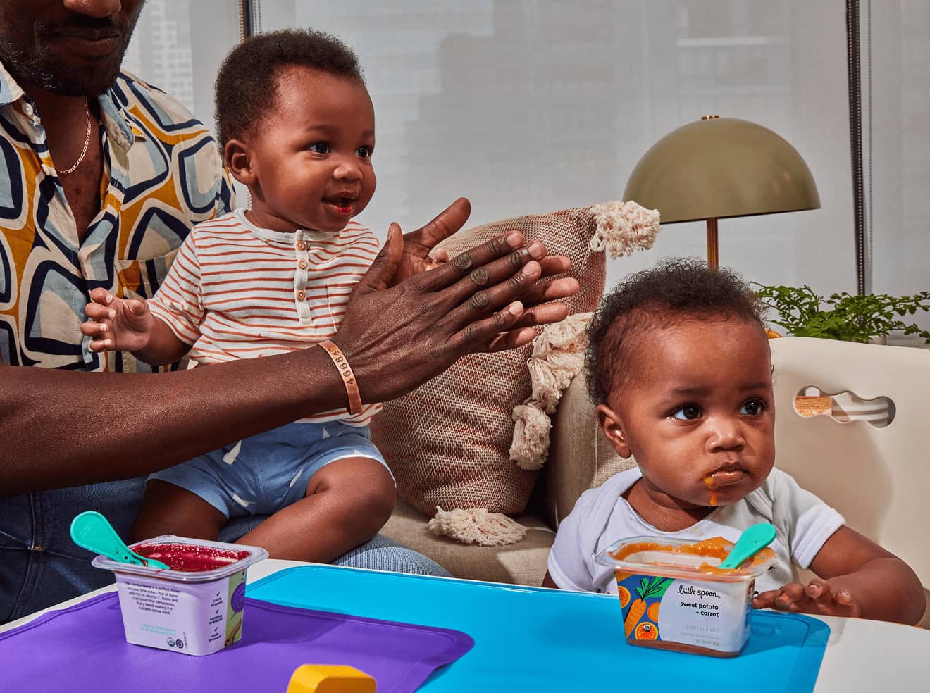 Two babies, one smiling while sitting on his dad’s lap while the other is standing while eating a 'sweet potato + carrot' Babyblends. The setting features a cozy living room with a lamp and cushions in the background.