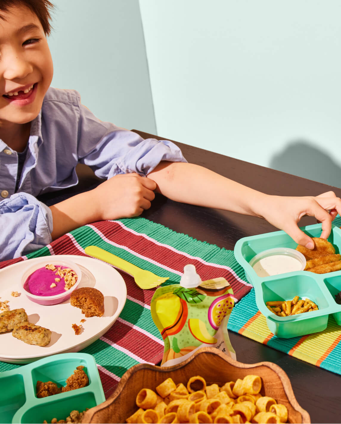 A child grabs on to a Chicken Super Nugget from the Chicken Dunkers luncher, while having the Brunch Lunch luncher on a plate, some Veggie Loops and an opened Smoothie served in front of them.