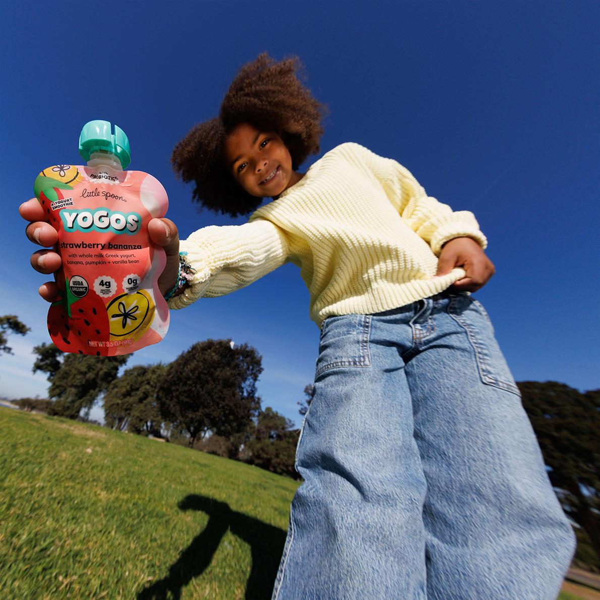 young girl with jeans and a sweater holding a Strawberry Bananza YoGo pouch in one hand standing in a park
