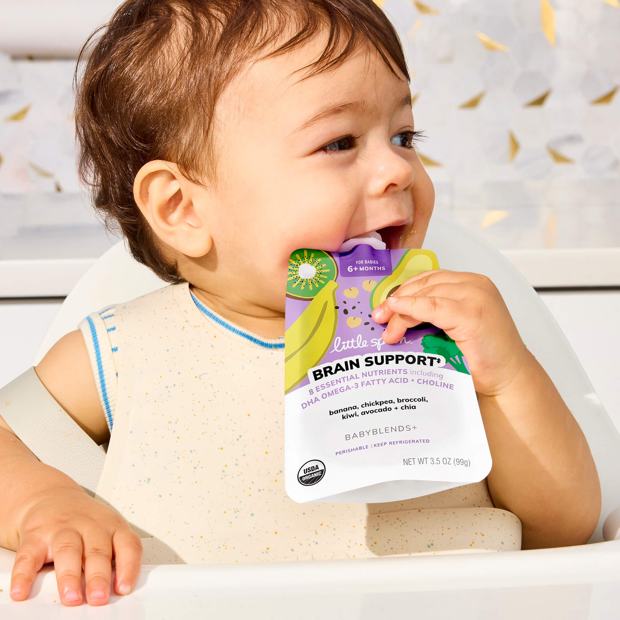 A baby sitting in a high chair holding a Banana, Chickpea, Broccoli, Kiwi, Avocado + Chia Babyblends+ pouch in its mouth