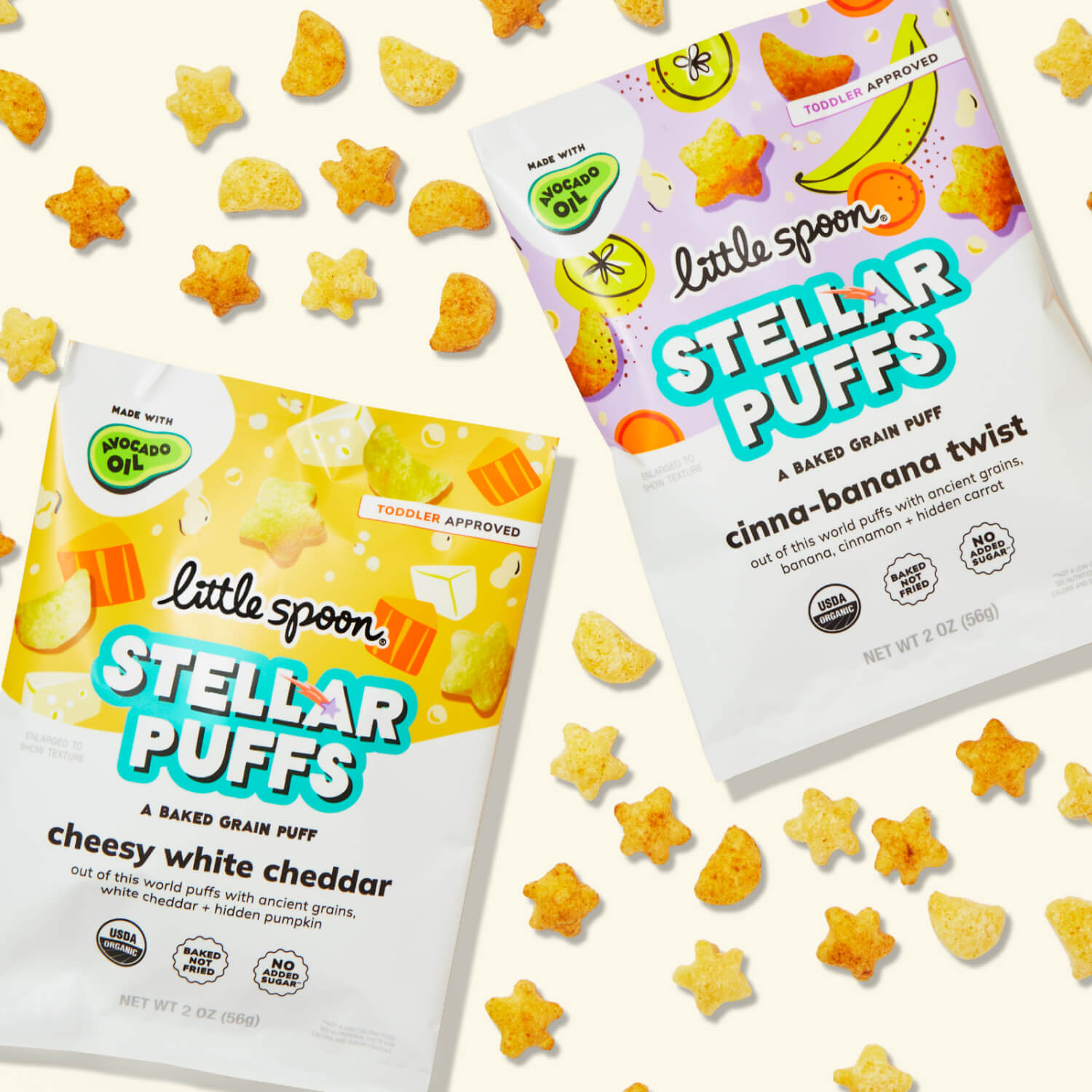 Sealed bags of Little Spoon Cheesy White Cheddar & Cinna-Banana Twist Stellar Puffs surrounded by star & moon shaped puffs.