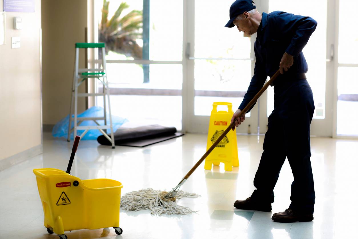 Here is a senior adult man working as a janitor in an office building, school or church.  He wears a uniform as he mops the entry floor.  Mop bucket and safety sign are also pictured.  He has retired and enjoys being active and still meeting people without responsibilities of the busy office demands.  Full length. header image