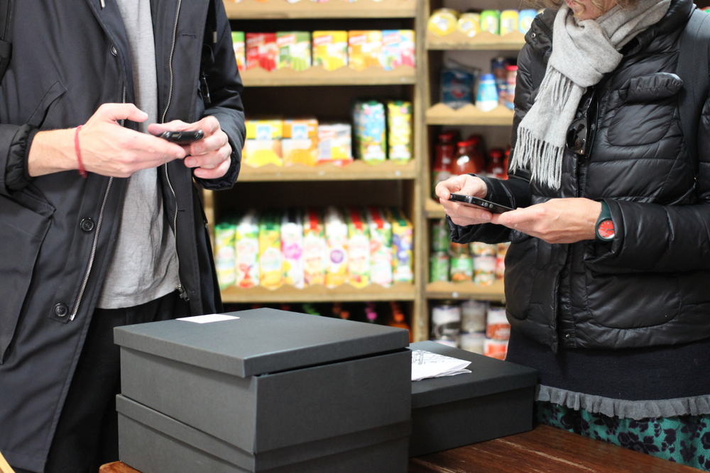 A cropped photo of a man and woman looking at their phones in front of a pile of black boxes with a white sticker on