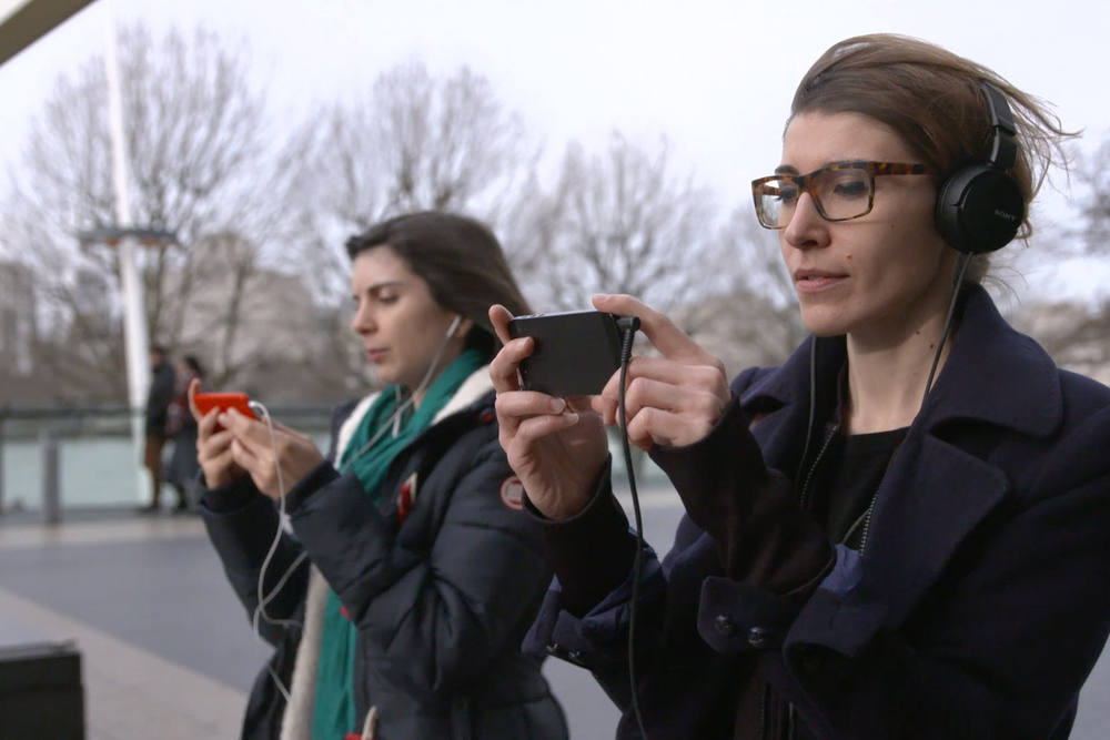 Two women with headphones on walking through London looking at a video playing on their phones that gives them directions