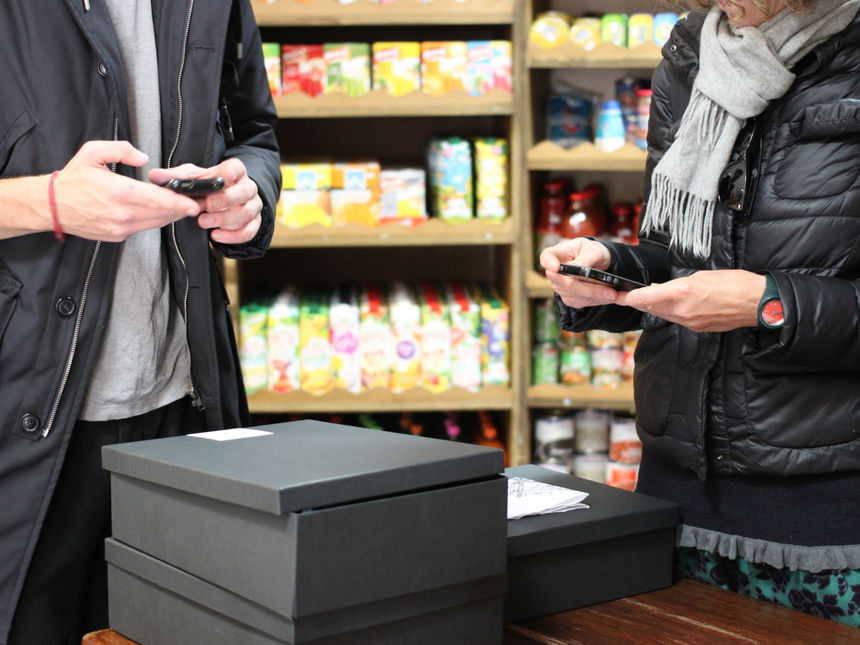 A cropped photo of a man and woman looking at their phones in front of a pile of black boxes with a white sticker on