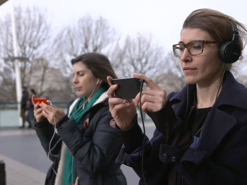 Two women with headphones on walking through London looking at a video playing on their phones that gives them directions