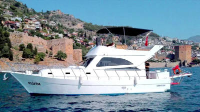 Elifim yacht cruising near a historic coastal town with hillside houses and castle walls in the background.