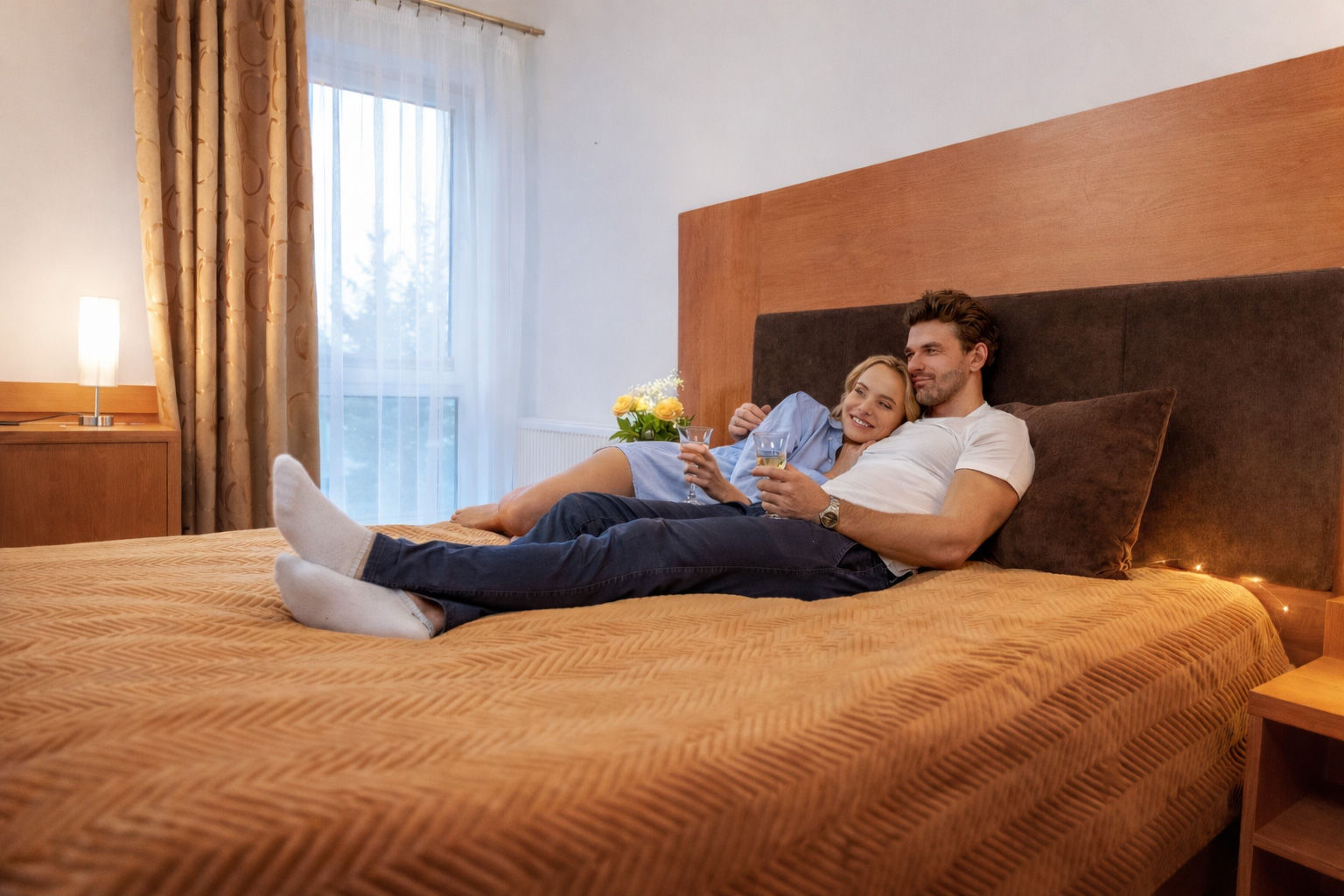 "Para wypoczywająca na łóżku w hotelowym pokoju z kieliszkami szampana, przytulna atmosfera w przytulnym wnętrzu hotelu PIASTUN."
