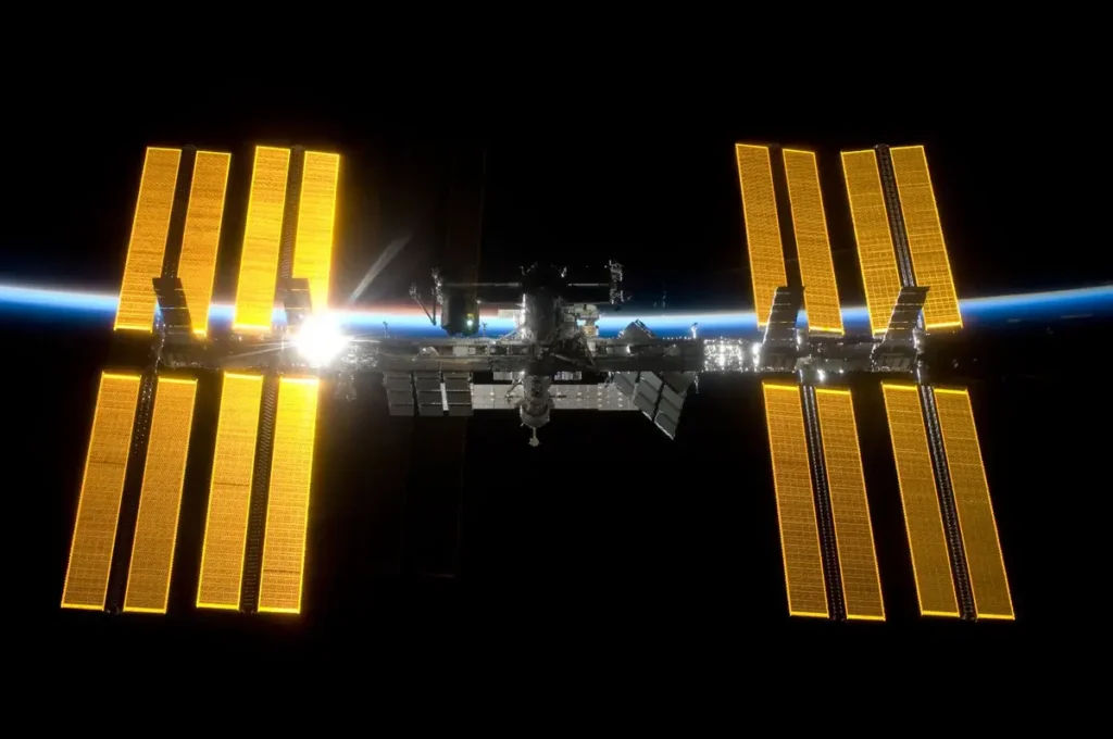 International Space Station with large solar panels orbiting Earth against the blackness of space and a distant moon.