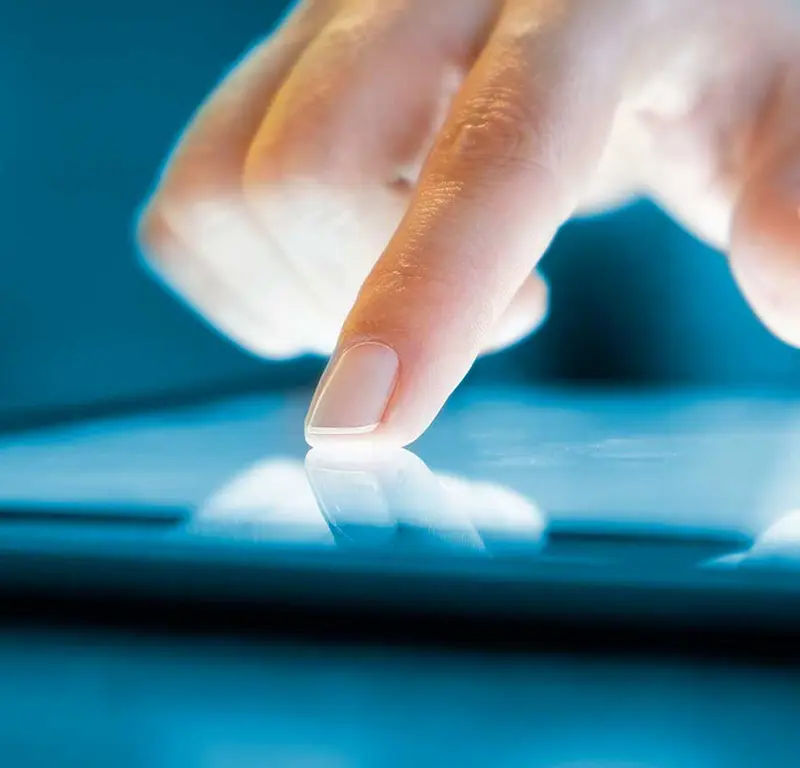Person in a blue shirt using a tablet with a finger tapping its screen, reflecting on the device glass.