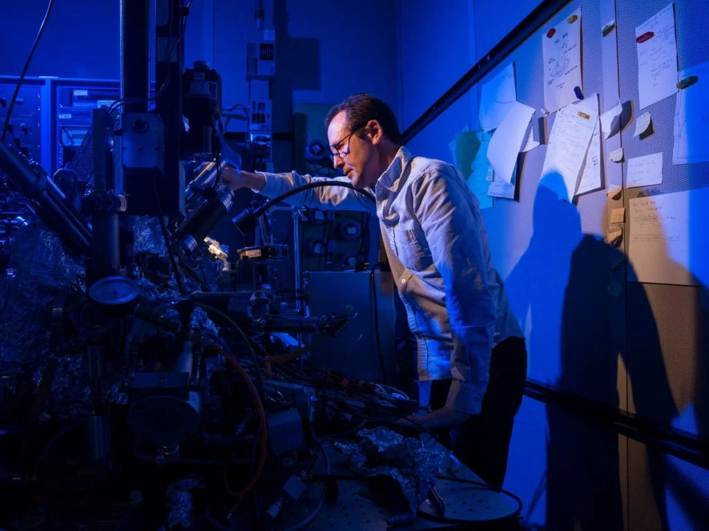 Scientist operates advanced machinery in a dimly lit lab with blue lighting and notes on the wall behind.