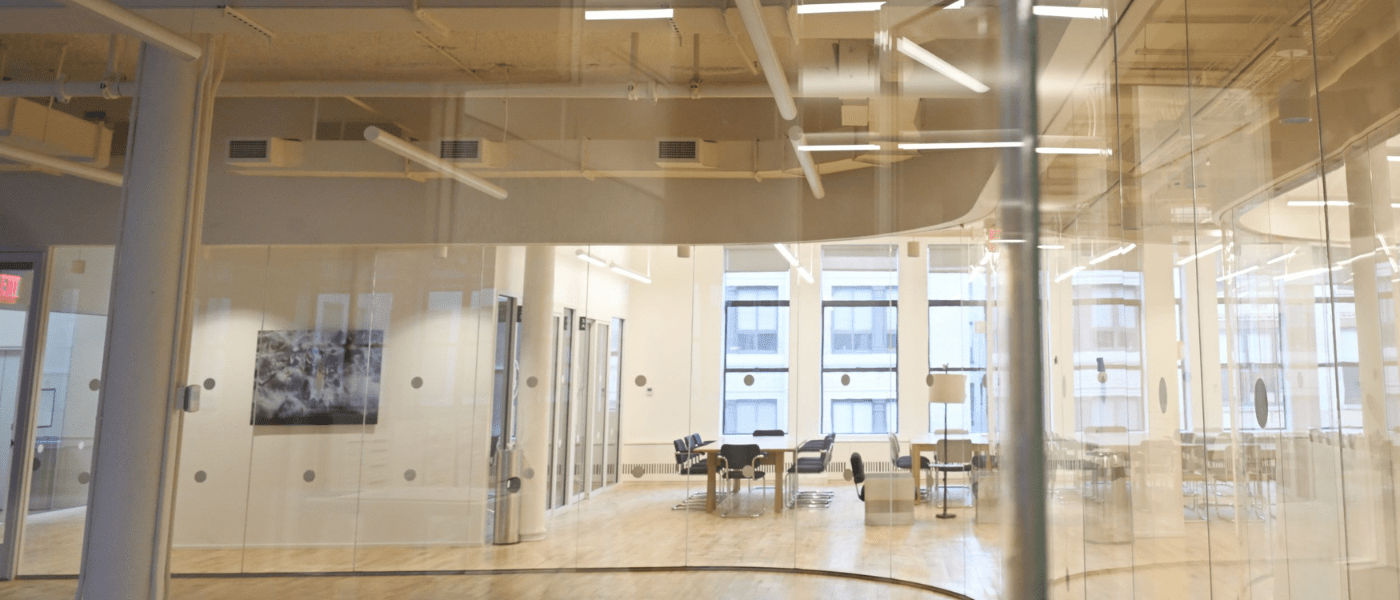 Modern office space with glass walls, wooden floors, tables, and chairs in a bright, open layout.