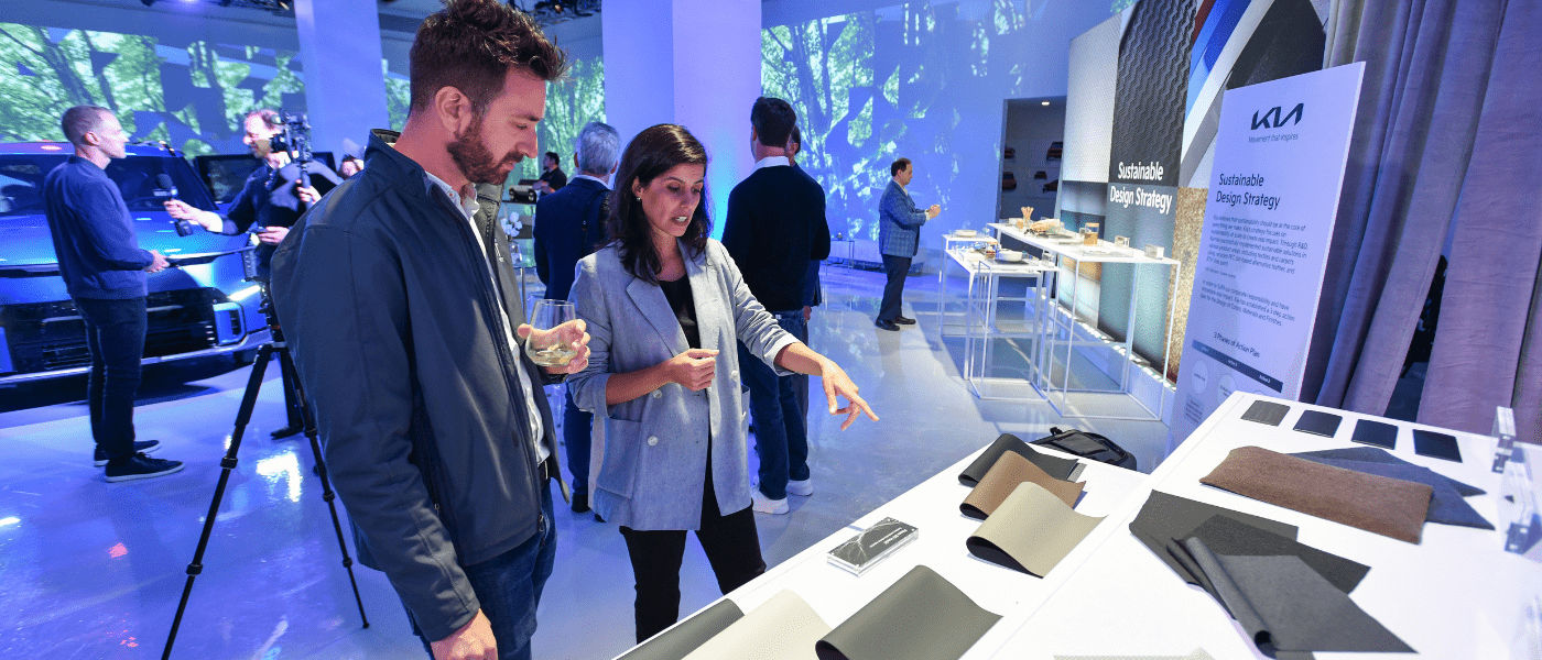A woman shows fabric samples to a man at a modern event with people and cars in the background.
