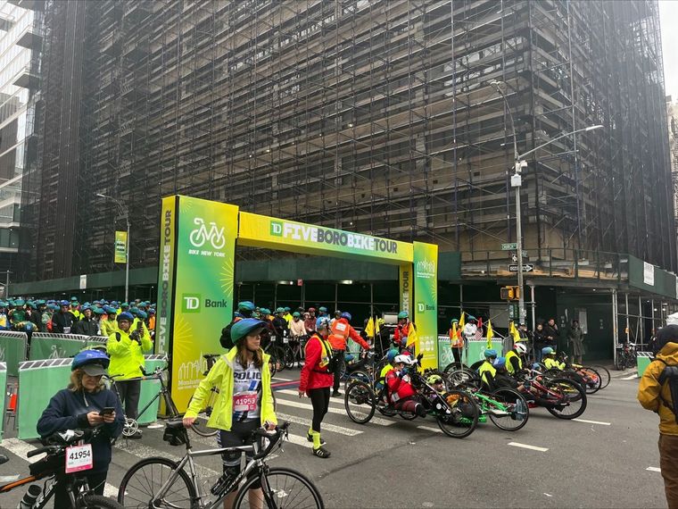 Cyclists gather near the starting line of the TD Five Boro Bike Tour in a city street with scaffolding.