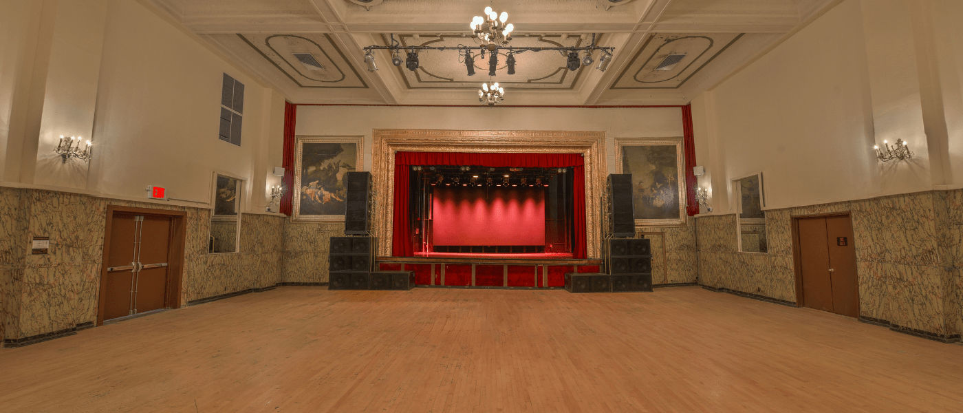An empty ballroom with a stage, chandeliers, and marble-paneled walls.