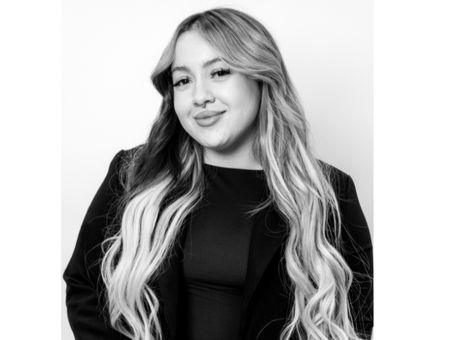 Woman with long wavy hair smiling, wearing a dark blazer, in front of a plain light background.