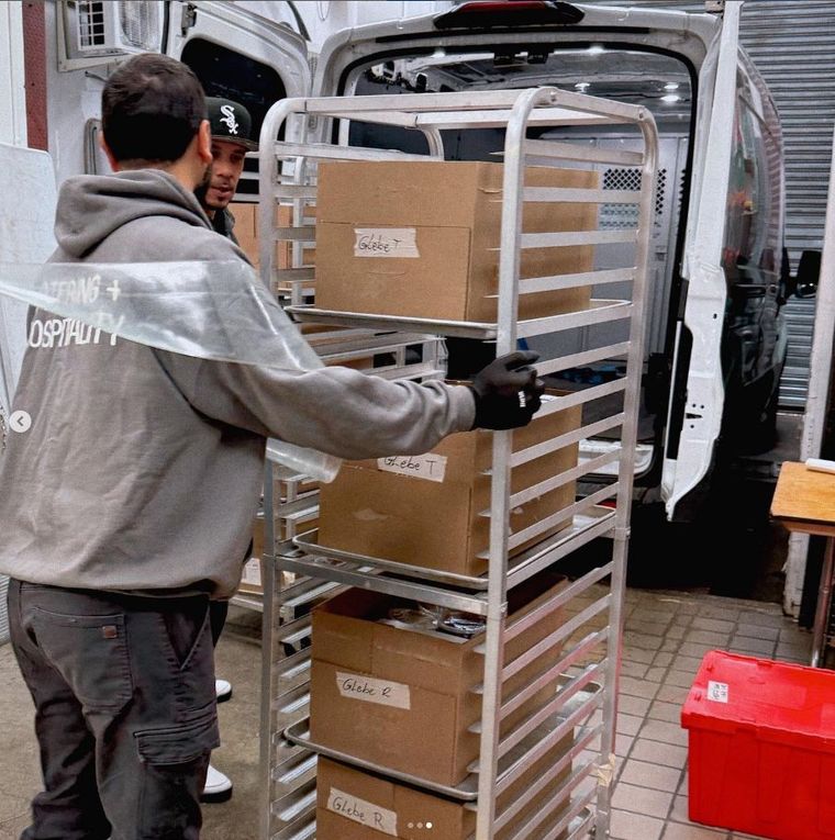 Two people load a metal rack with labeled boxes into a white delivery van at a loading area.