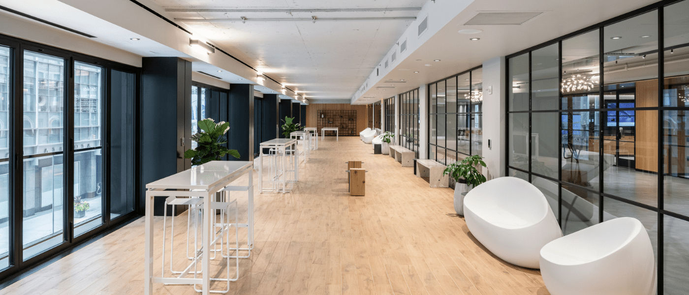 Modern open office space with large windows, white furniture, wood floor, and glass-walled meeting rooms.