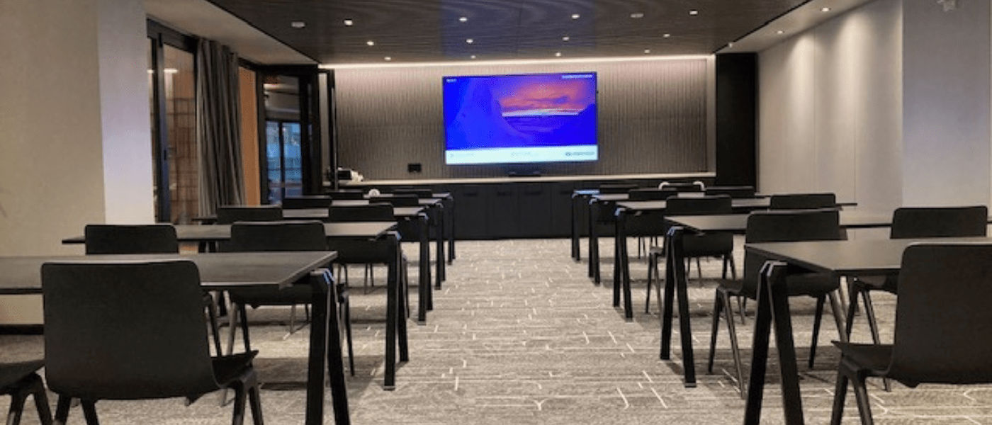A modern conference room with rows of black tables and chairs facing a large screen at the front.