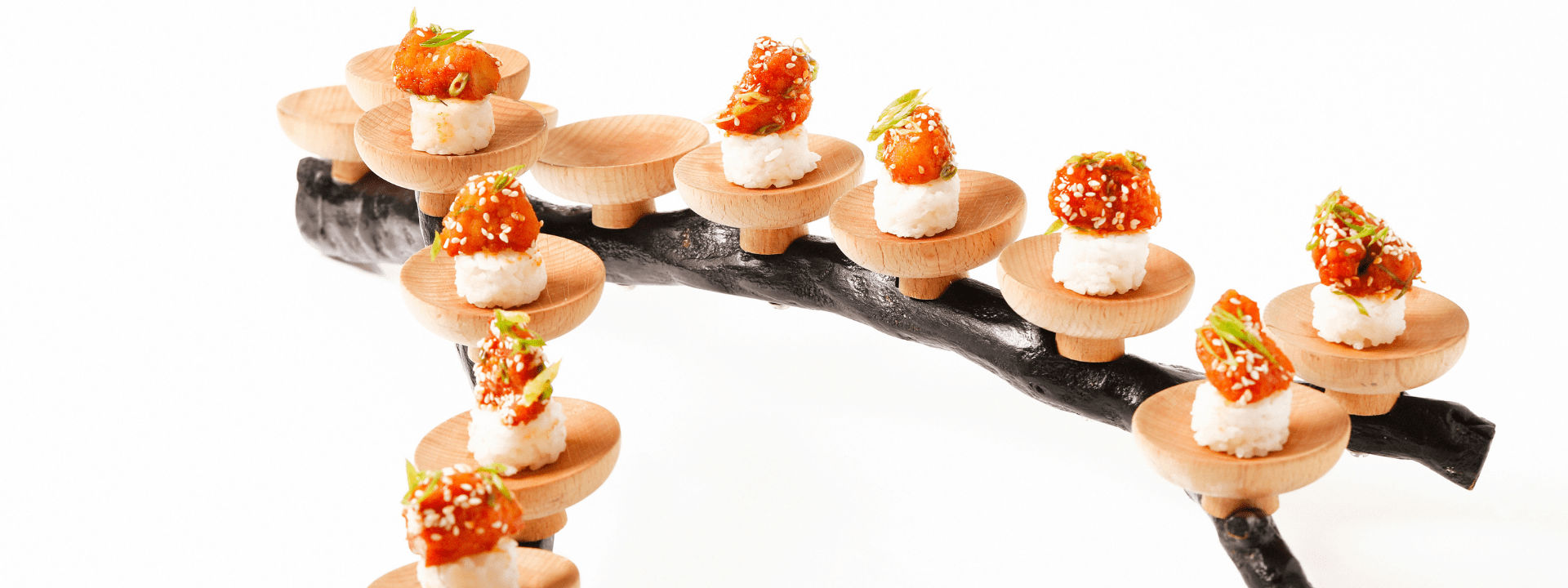 Small sushi pieces topped with fish, arranged on wooden stands atop a black decorative branch.