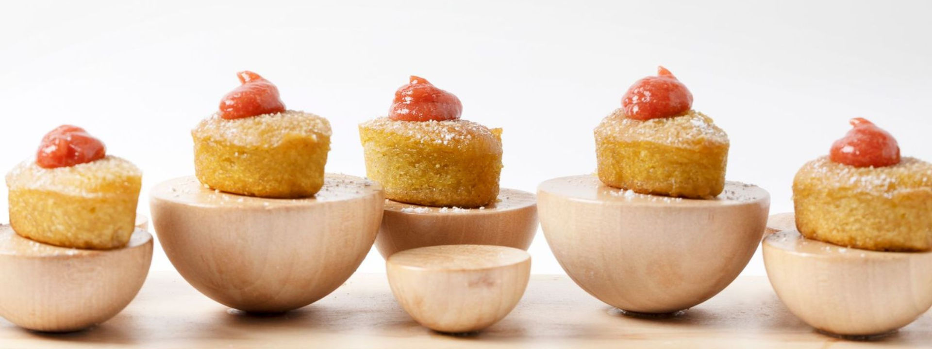Small golden cakes topped with red sauce, displayed on round wooden stands against a white background.