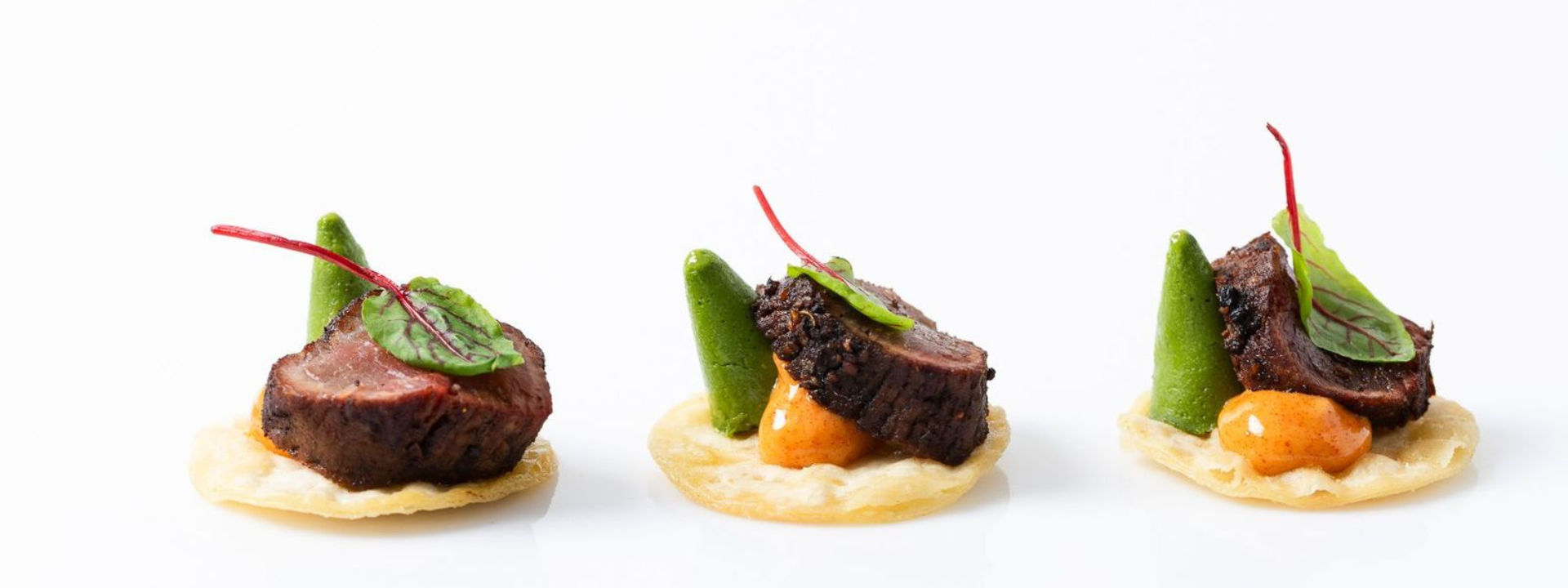 Three gourmet appetizers with seared meat, green puree, leaf garnish, and sauce on round crispy bases.