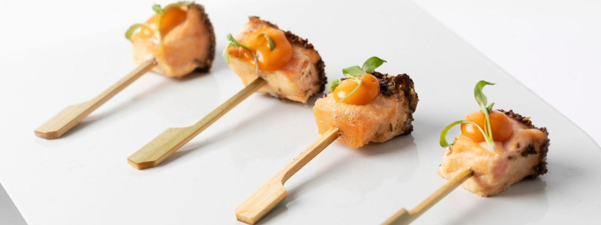 Four bite-sized salmon skewers garnished with orange sauce and green herbs on a white plate.