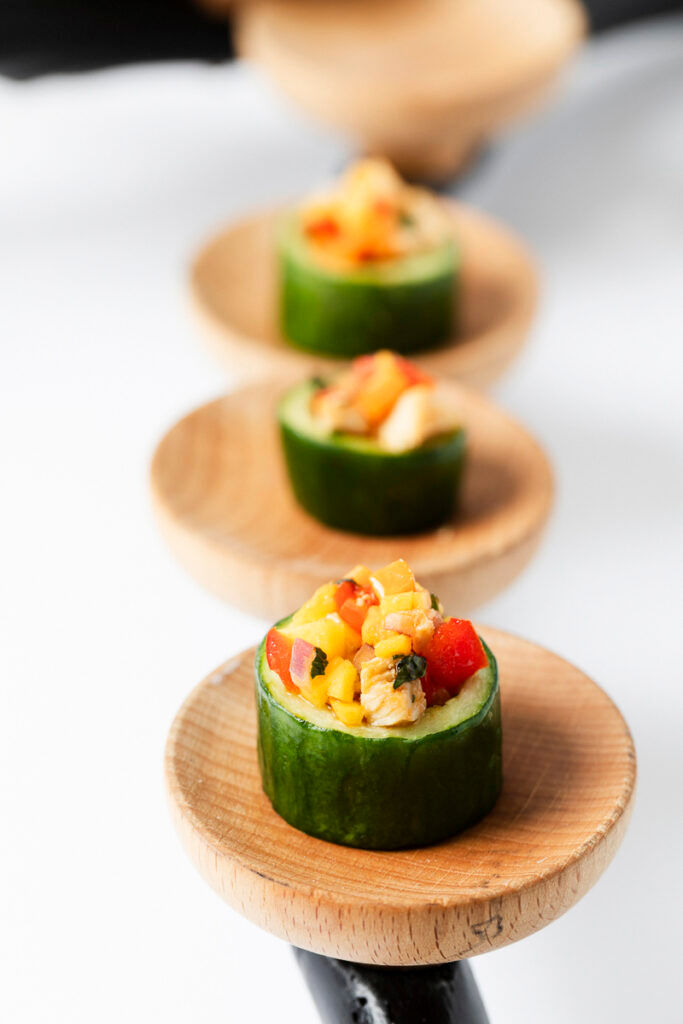 Cucumber rounds topped with colorful diced vegetables, served on small wooden plates in a row.
