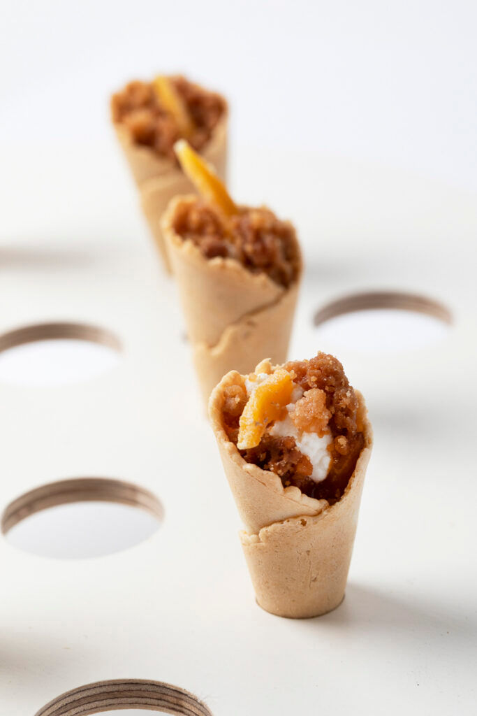 Mini dessert cones filled with crumble and cream, arranged in a row on a white surface with circular holes.