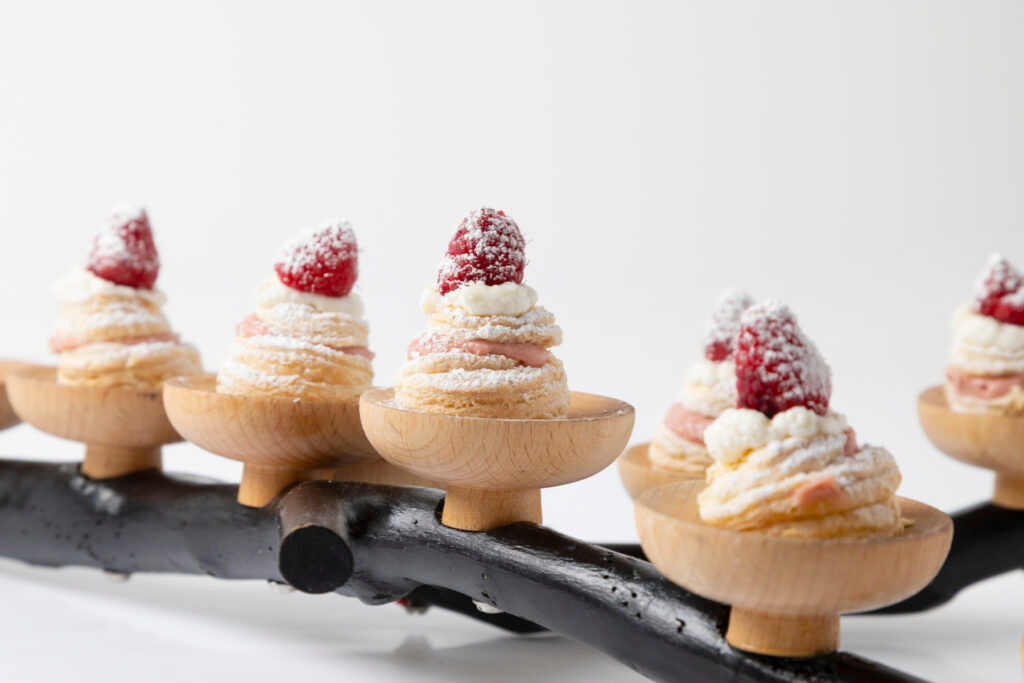Mini cream puffs topped with raspberries and powdered sugar, served on wooden stands over a dark branch.