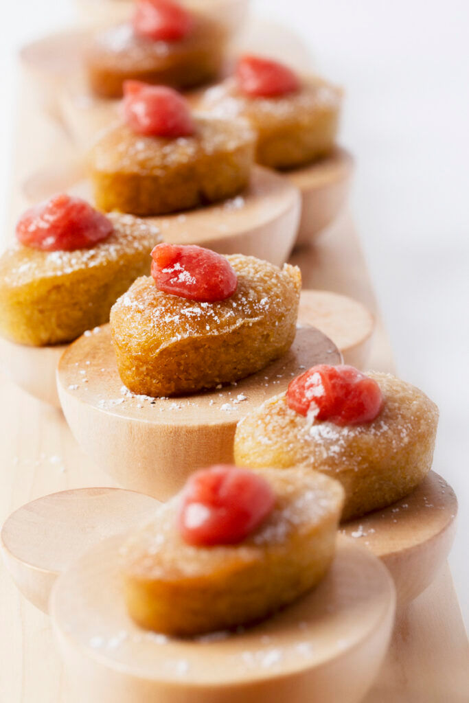 Small golden cakes topped with red sauce and powdered sugar, arranged neatly on round wooden pedestals.