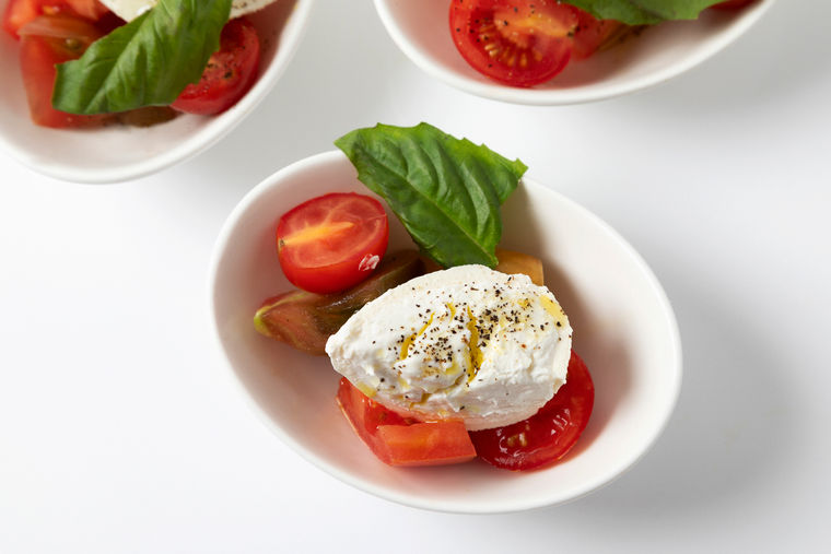 Small white bowl with burrata cheese, sliced tomatoes, basil leaf, olive oil, and black pepper.