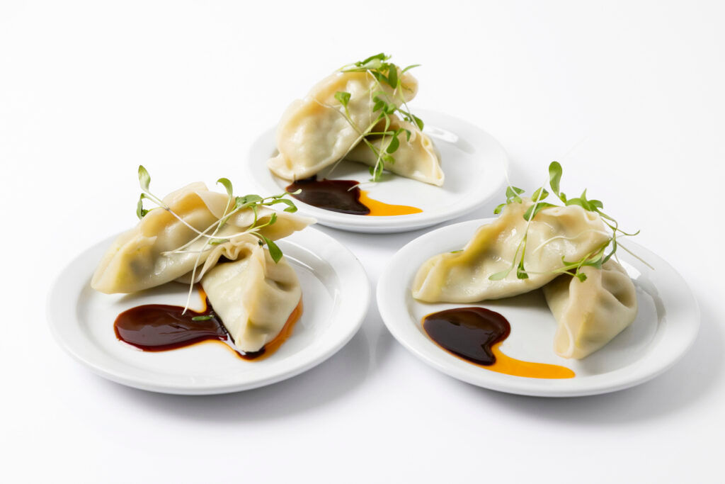 Three plates with dumplings, dark and orange sauce, and microgreens on a white background.