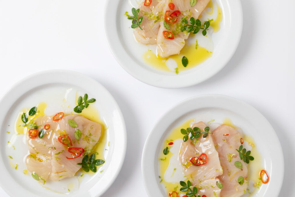 Three white plates with fish crudo, olive oil, sliced red chili, herbs, and zest on a white background.
