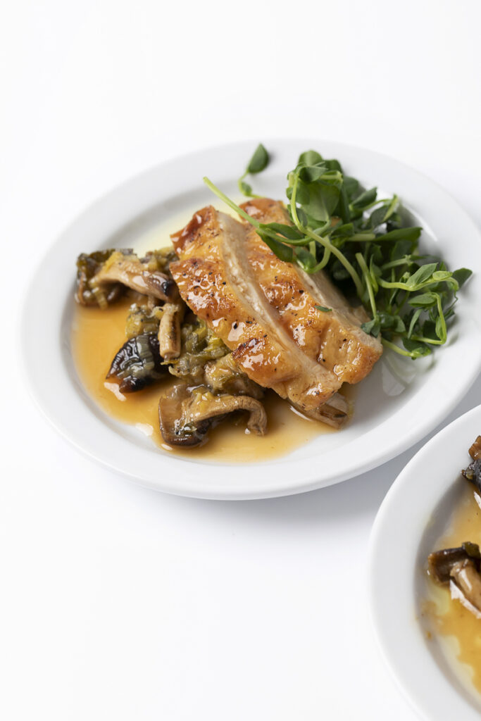 A plate with glazed roasted chicken, sautéed mushrooms, greens, and sauce on a white background.