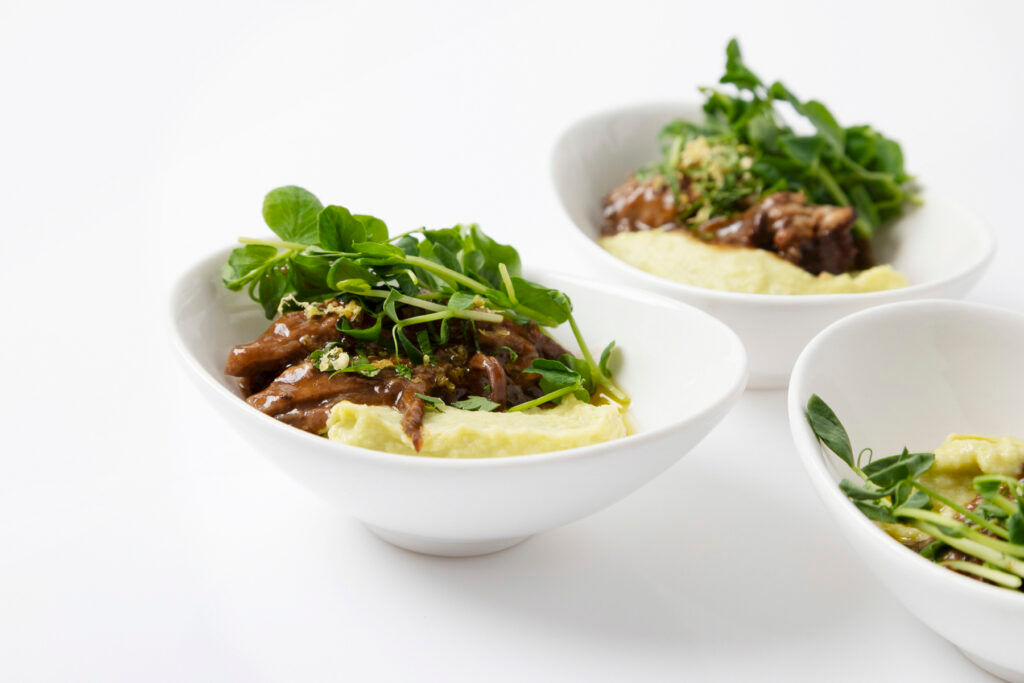 Three white bowls with mashed potatoes, beef stew, and fresh green herbs on a white background.