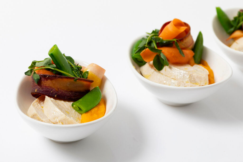Small white bowls with sliced chicken, colorful carrot ribbons, snap peas, and greens on a white background.