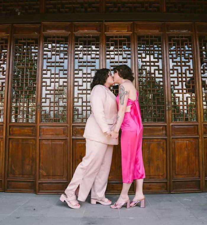 Two people holding hands and kissing in front of a wooden building with ornate window panels.