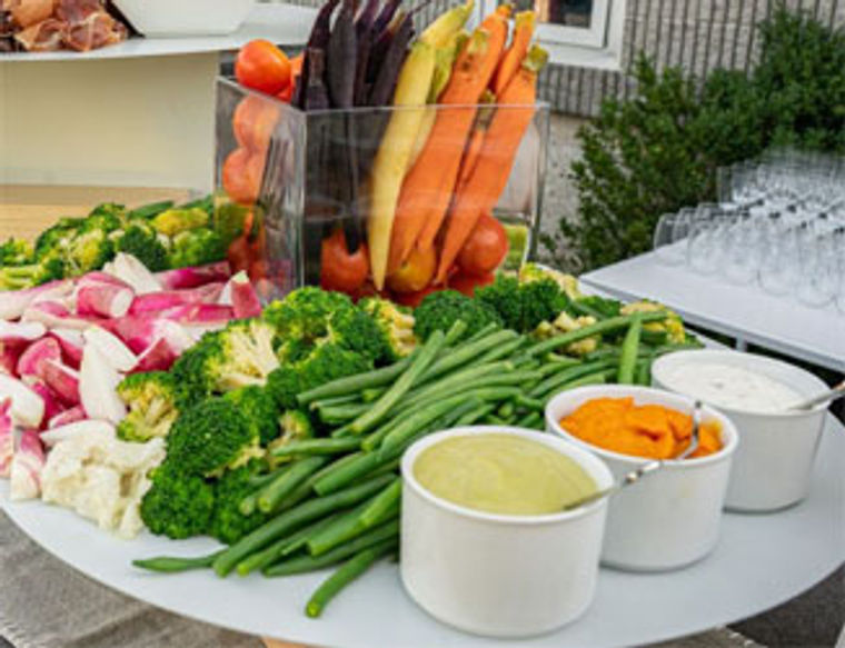 A platter of fresh vegetables with three dips in bowls on a table outdoors.
